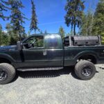 Ridgeline rooftop tent mounted on an overland pod truck bed rack system with rounded bars and lockable storage boxes on a black pickup truck, parked outdoors on Vancouver Island surrounded by tall trees and blue skies.
