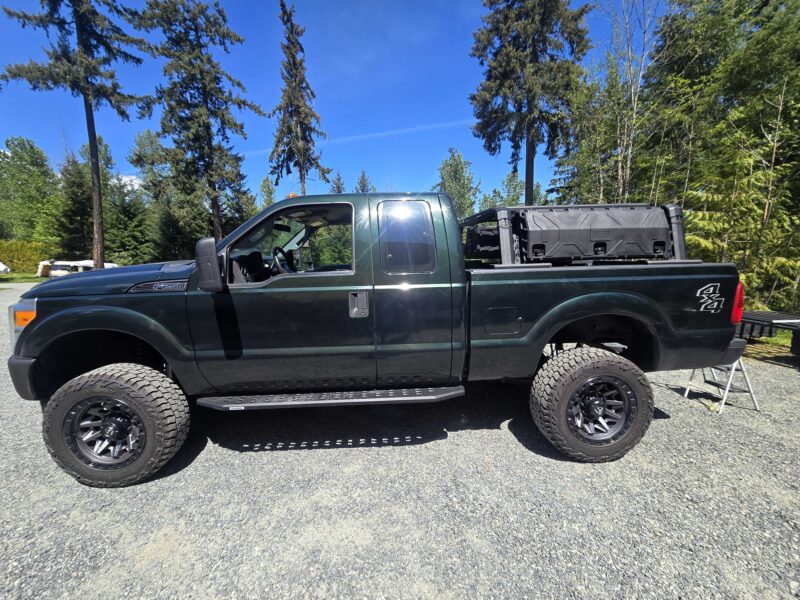 Ridgeline rooftop tent mounted on an overland pod truck bed rack system with rounded bars and lockable storage boxes on a black pickup truck, parked outdoors on Vancouver Island surrounded by tall trees and blue skies.